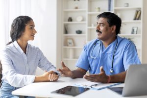 Indian Doctor In Uniform Consulting Smiling Female Patient During Meeting In Clinic